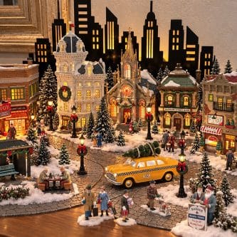 Elaborate miniature Christmas village with lit buildings, snow, trees, and a yellow taxi surrounded by tiny figures in a festive scene.
